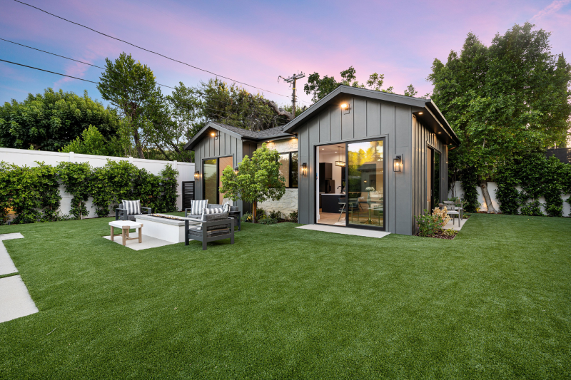 Modern gray house with large windows, surrounded by artificial grass lawn, outdoor seating area, and trees under a pink and blue sky at sunset.